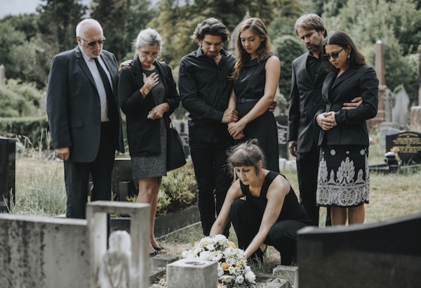 Mourners dressed in black gather at a cemetery, placing flowers on a grave.